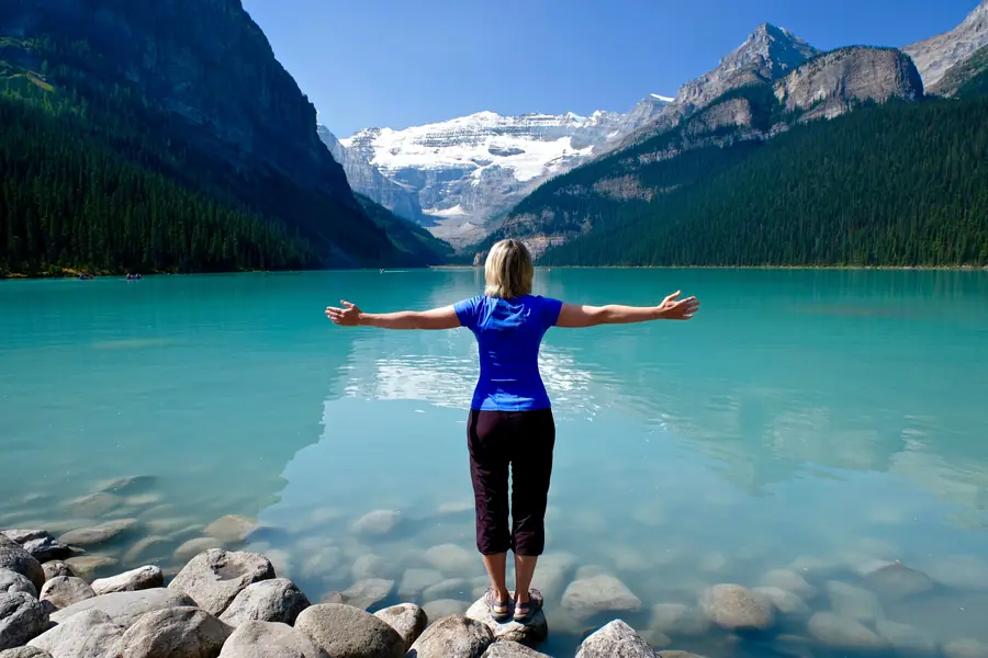 A woman standing on rocks at the edge of a turquoise mountain lake, arms open wide.