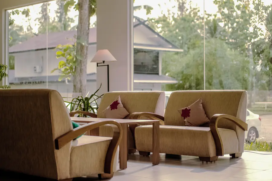 A bright, plant-filled waiting room with armchairs arranged around a low wooden table beside full-height windows.