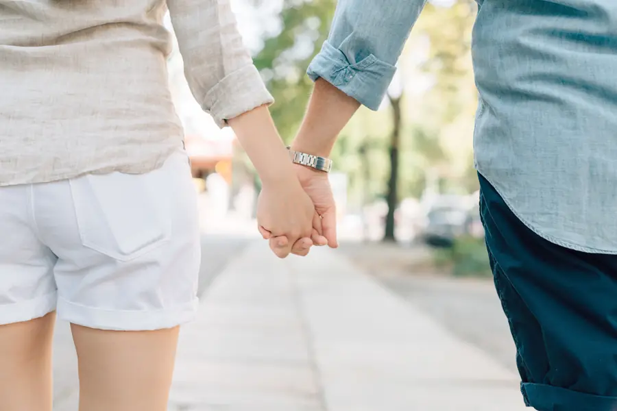 A couple walking along a tree-lined pavement, holding hands loosely.