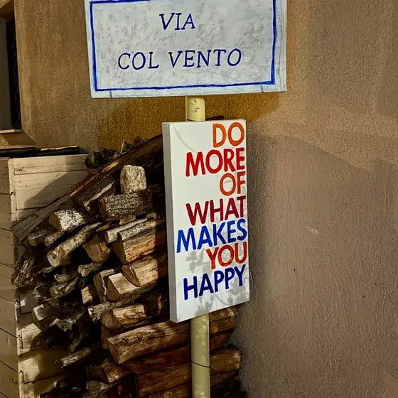 A street sign reading Via Col Vento beside a poster reading &ldquo;Do more of what makes you happy&rdquo;.